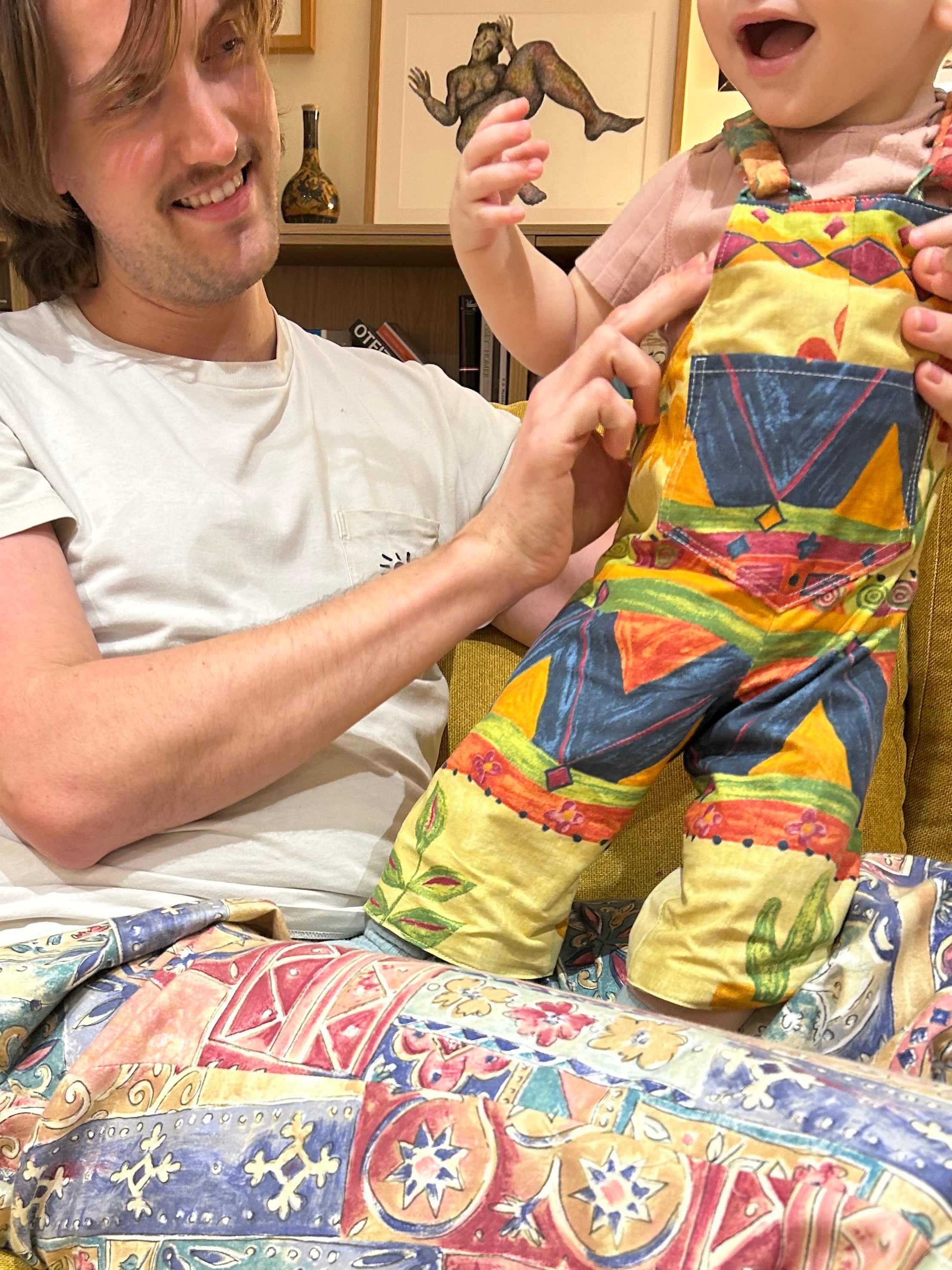 Man holding a child wearing colorful pants with geometric patterns in a home setting.