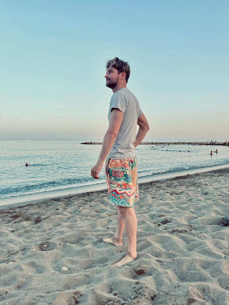 Man standing on a beach wearing colorful shorts and a light gray t-shirt.
