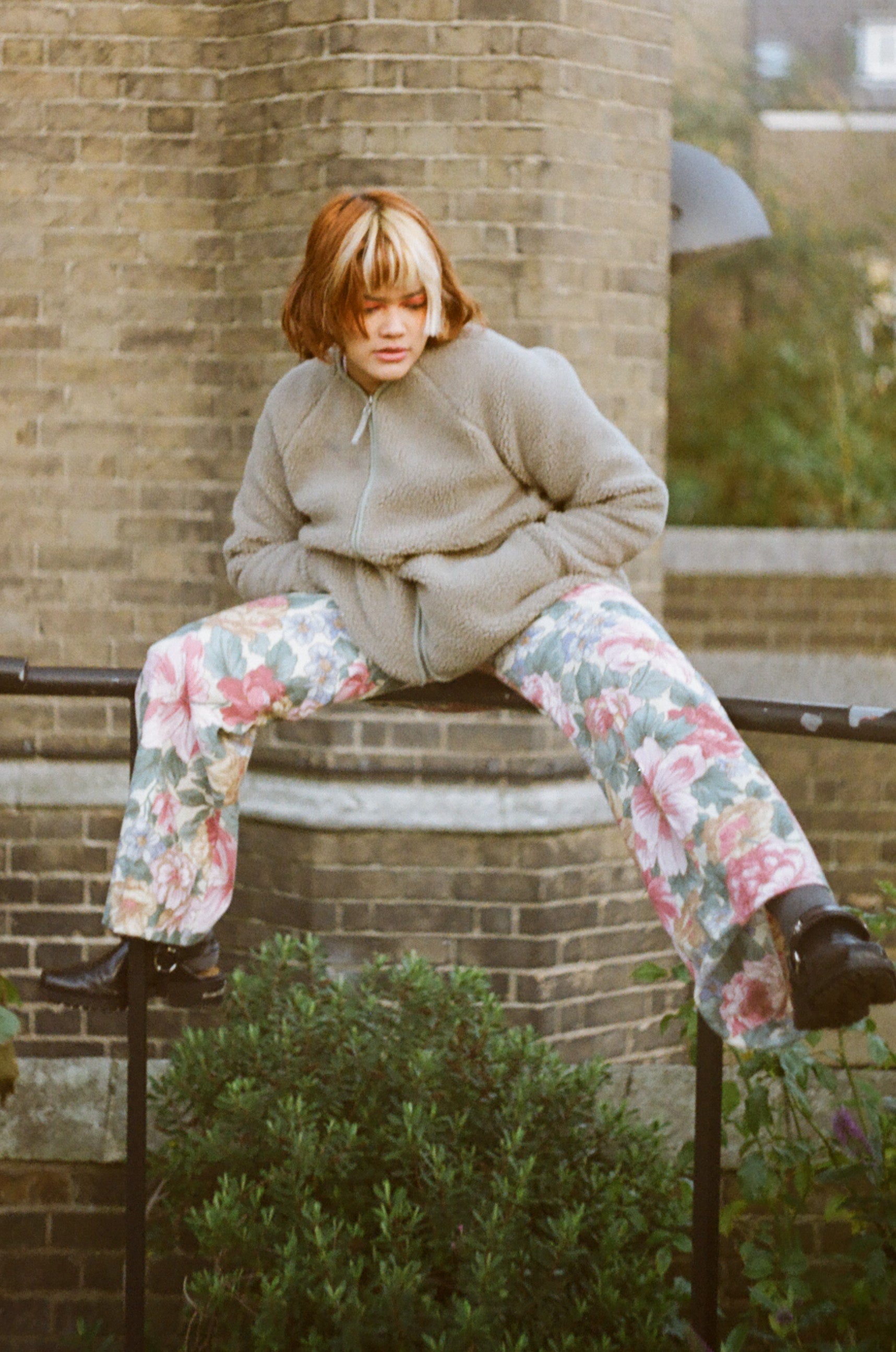 Person wearing a gray sweater and floral pants sitting on a ledge.