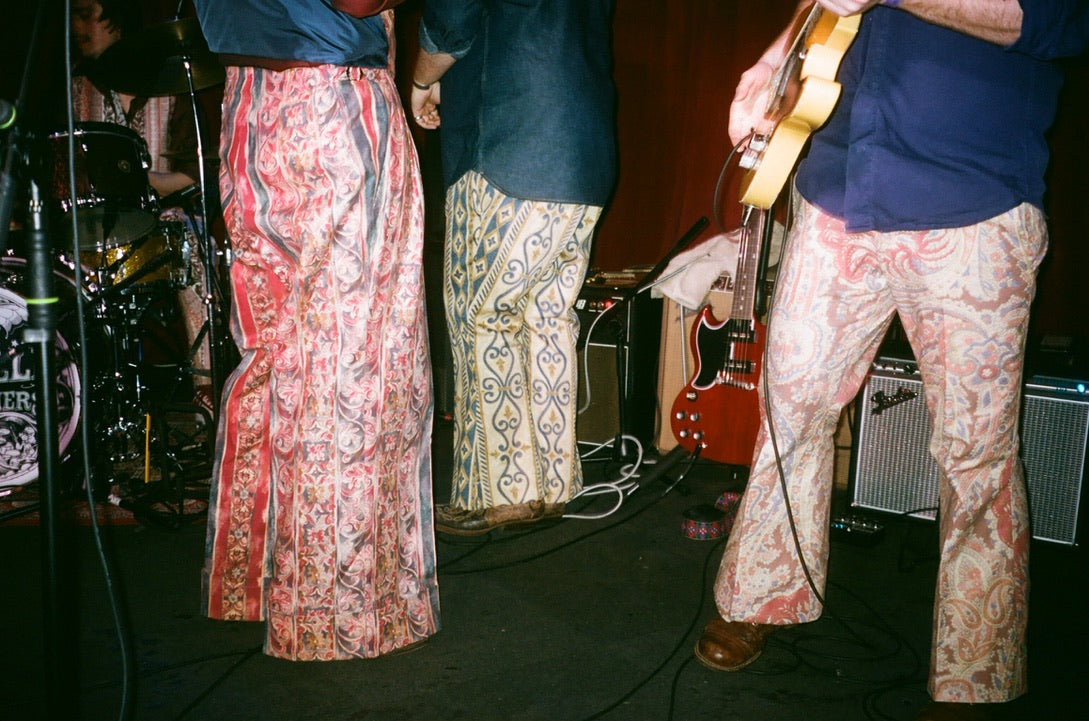 Three people on stage wearing patterned pants with musical equipment and a bottle in the background.