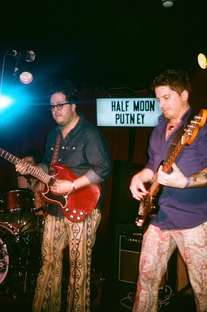 Two musicians playing guitar on stage with a 'Half Moon Putney' sign in the background.