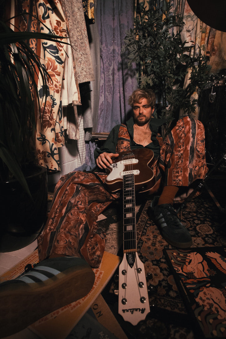 Man sitting with a guitar in a room with patterned walls and furniture