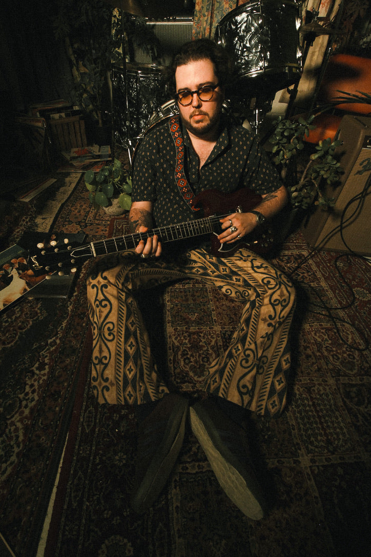 Man playing guitar in a dimly lit room with patterned rug and plants.