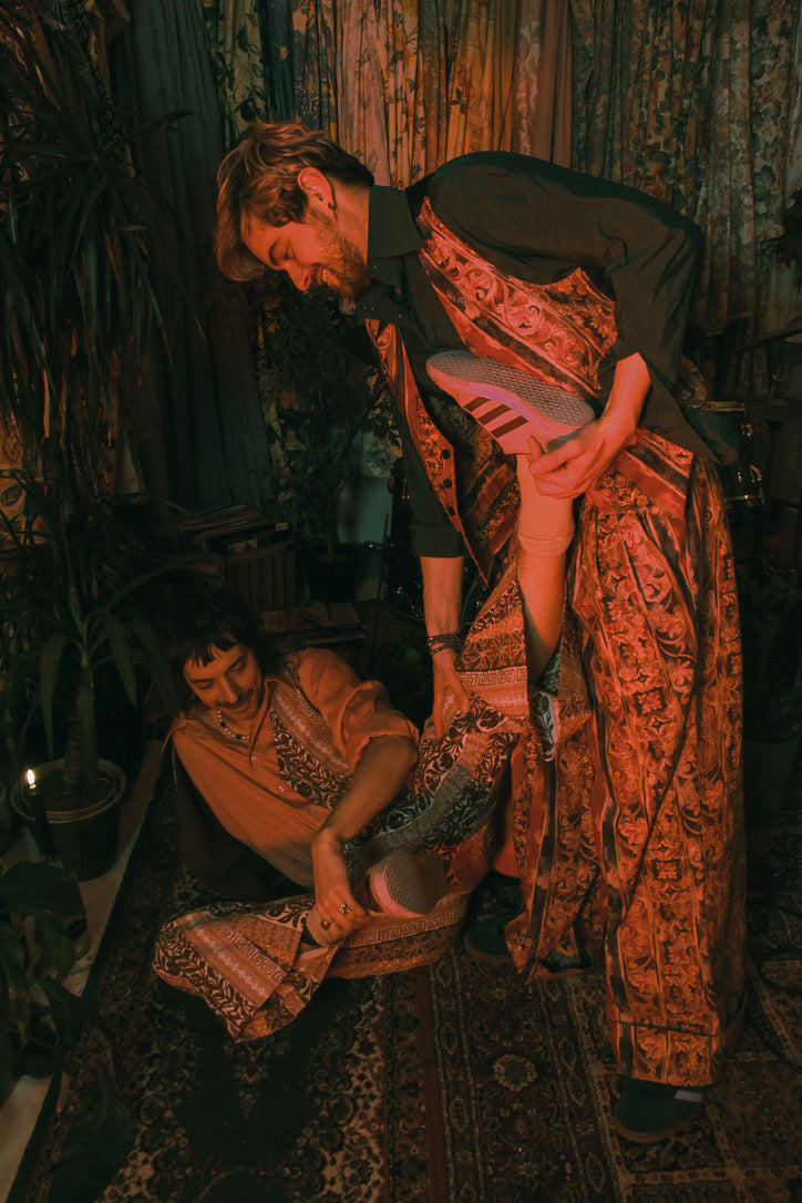 Two people wrapped in patterned cloths in a dimly lit room with plants.