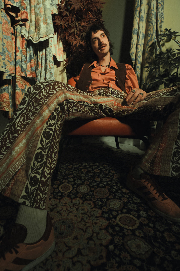 Person sitting on a chair with patterned pants and shirt in a room with floral curtains and rug.