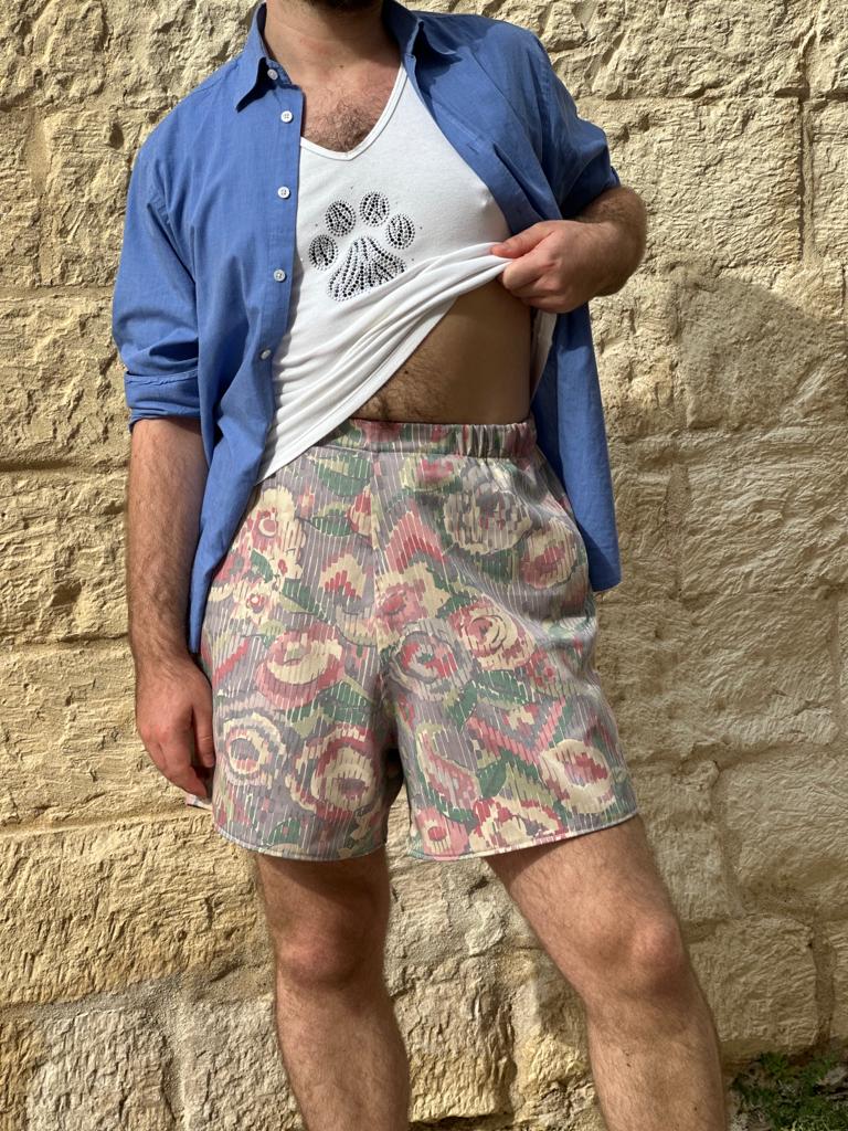 Person wearing a blue shirt over a white tank top and colorful patterned shorts against a stone wall.
