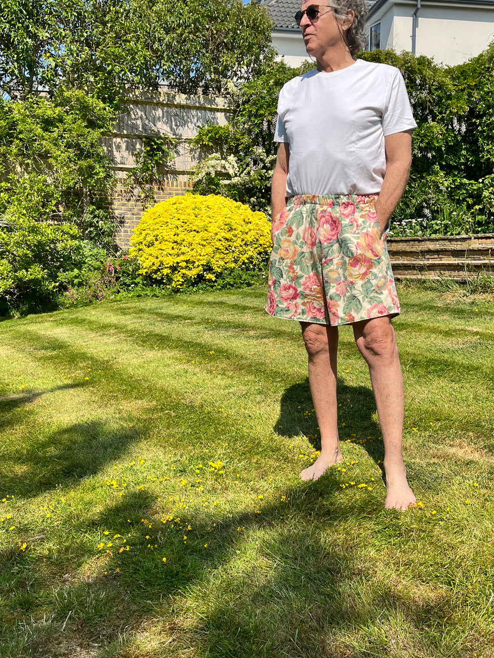 Person wearing a white t-shirt and floral shorts standing on grass with trees and a house in the background.