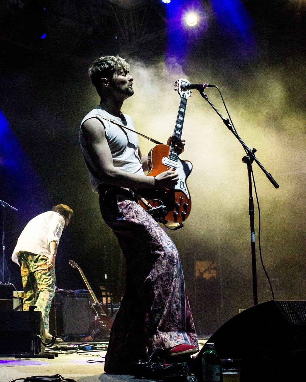 Person playing guitar on stage with a microphone stand and another person in the background.