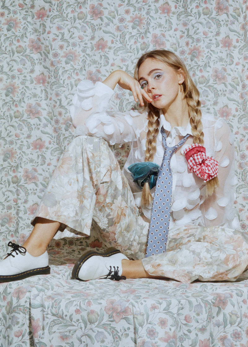 Young girl sitting on a floral-patterned couch wearing a white outfit with a tie and polka dot overalls.