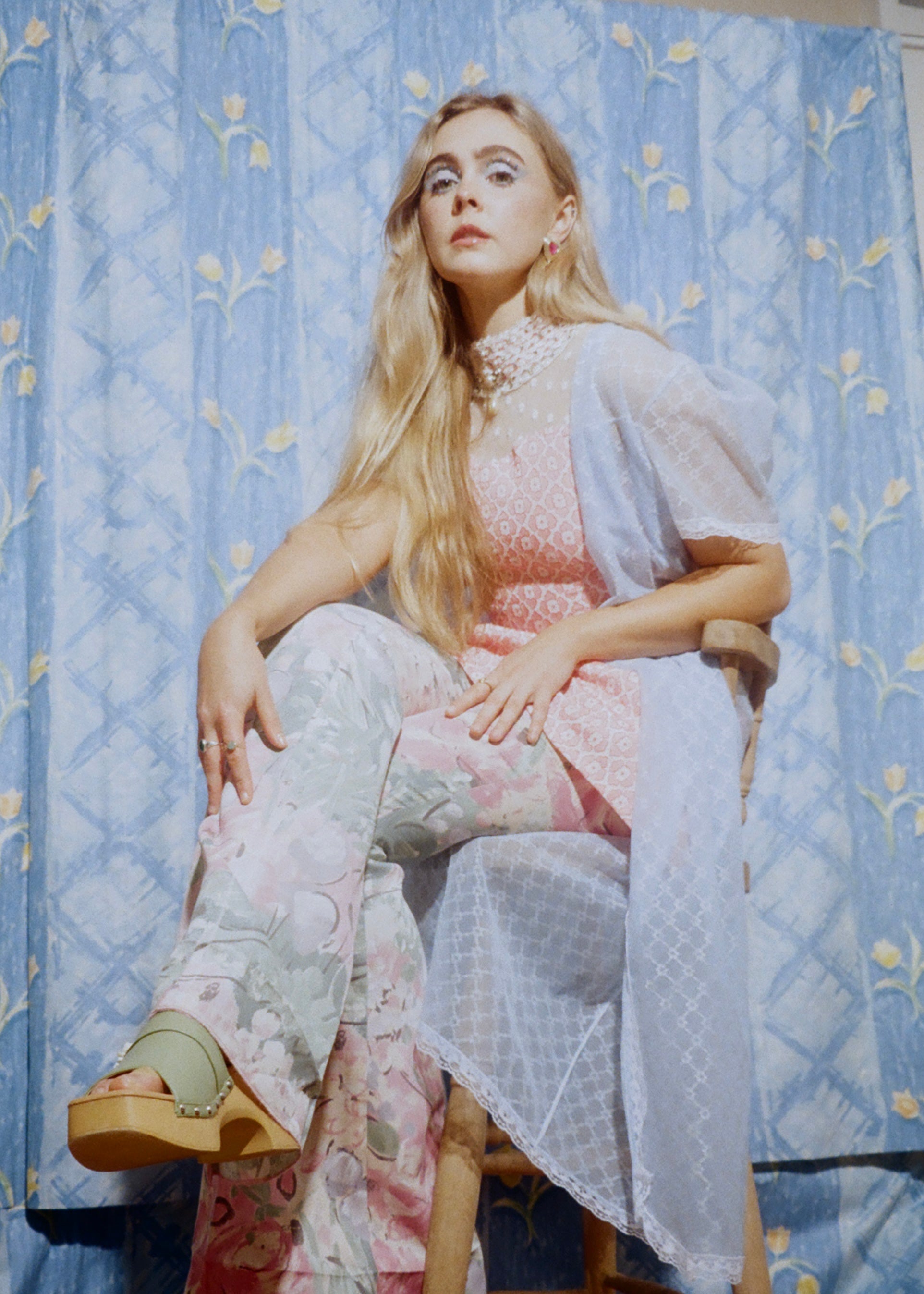 Woman sitting on a chair wearing a patterned dress against a decorative blue wall.