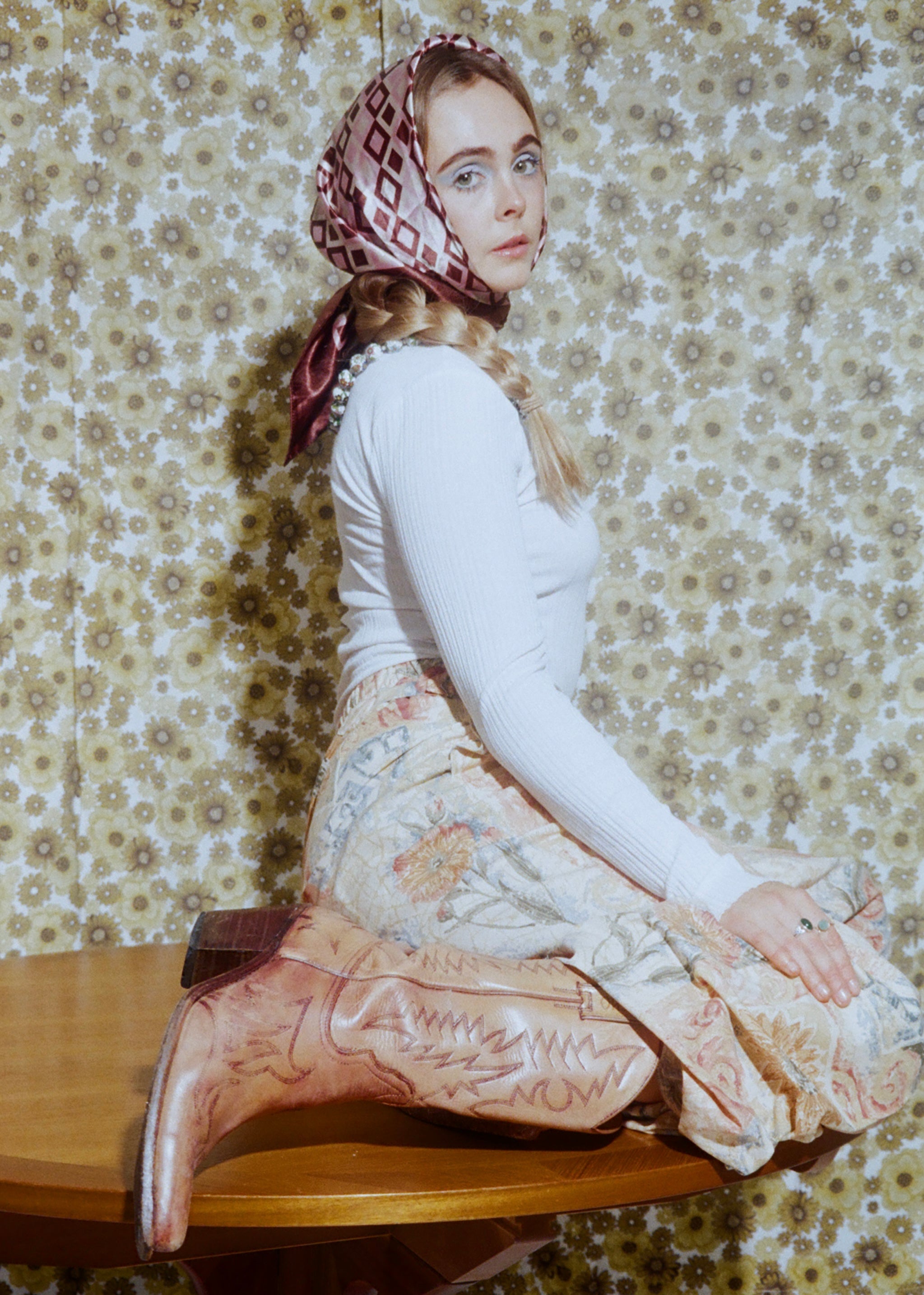 Woman sitting on a wooden stool wearing a patterned headscarf and floral skirt against a floral wallpaper background.