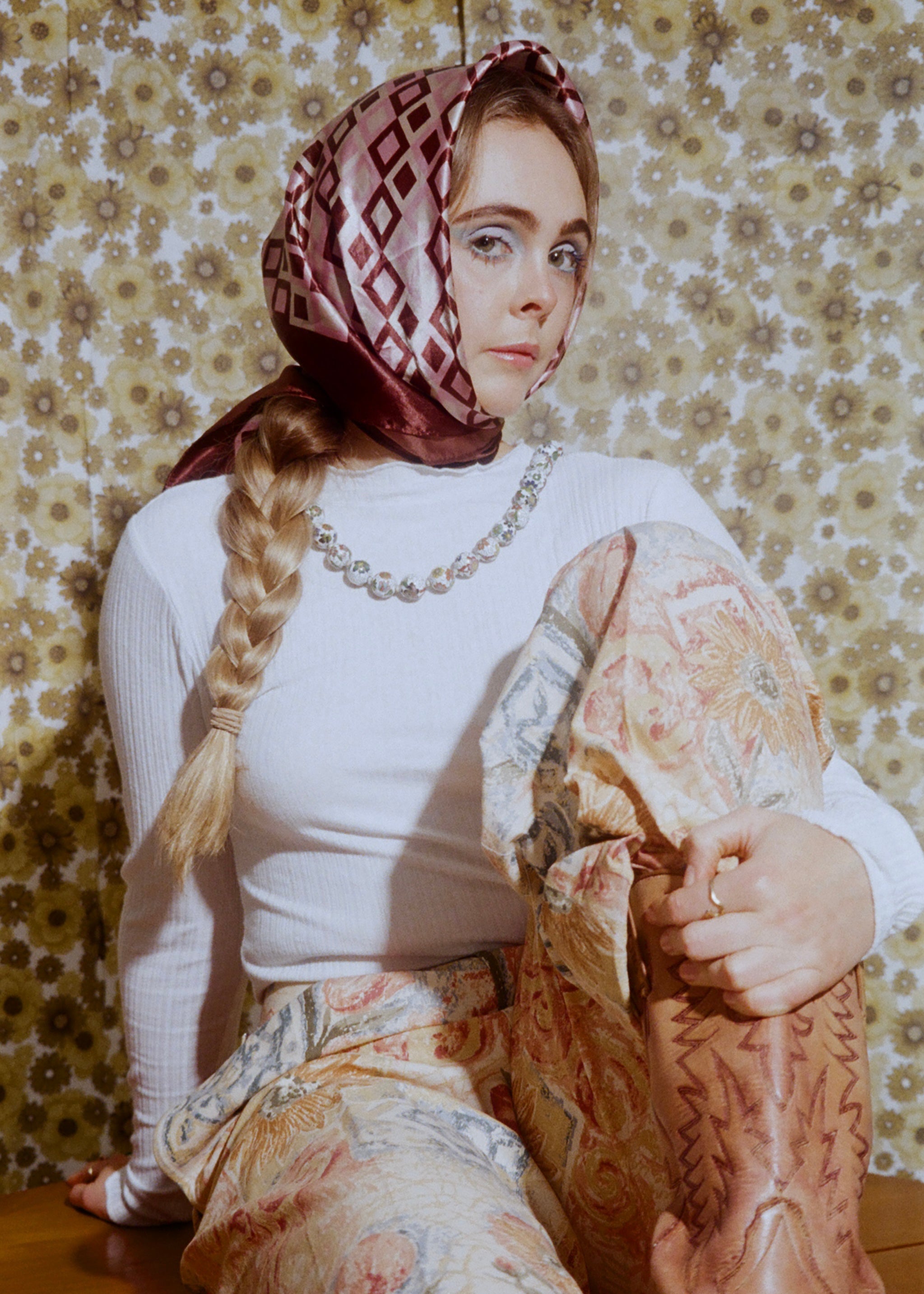 Woman wearing a patterned headscarf and sitting against a floral wallpapered wall.