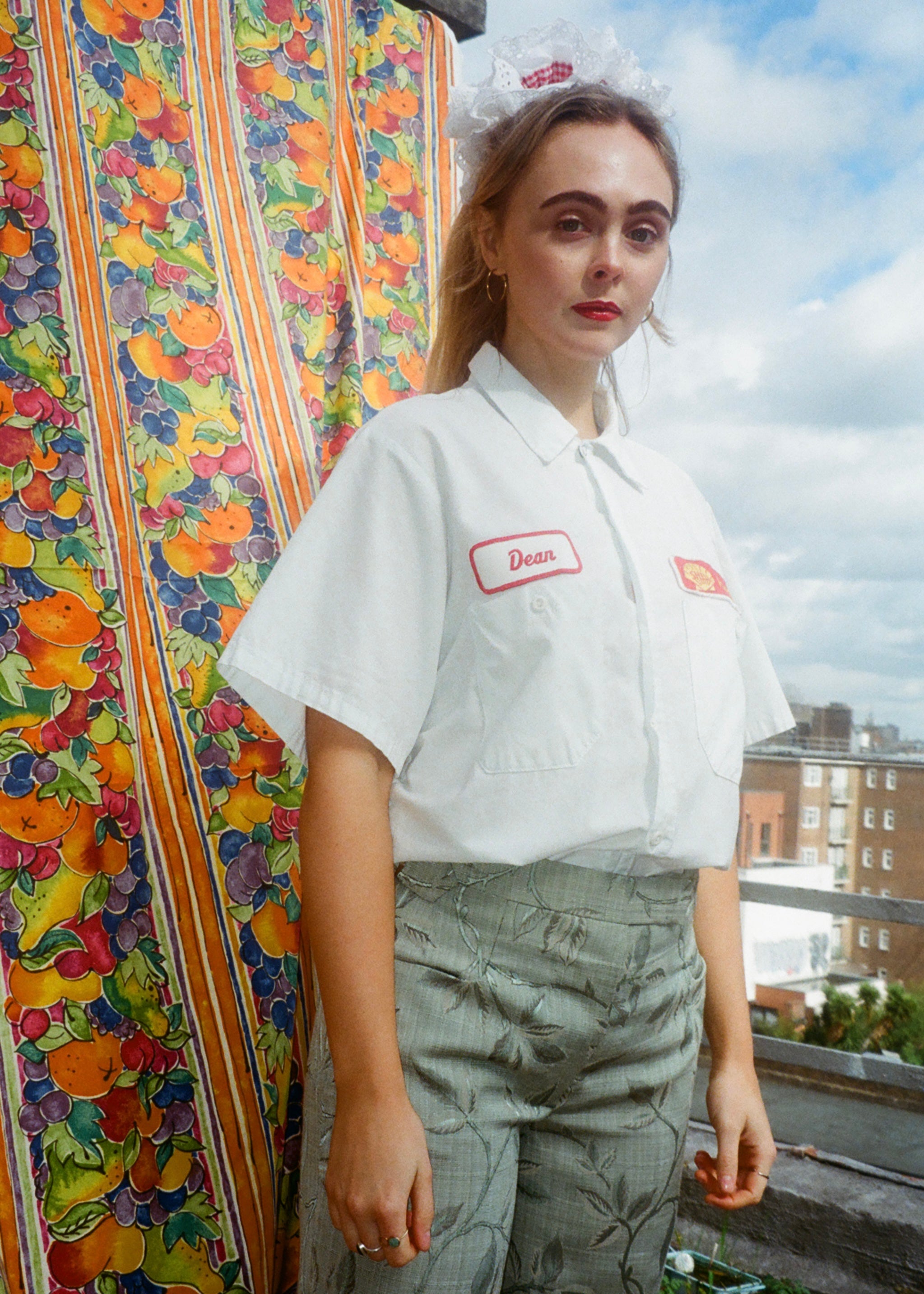 Person wearing a white shirt with a logo and green pants standing against a colorful fabric and cityscape background.