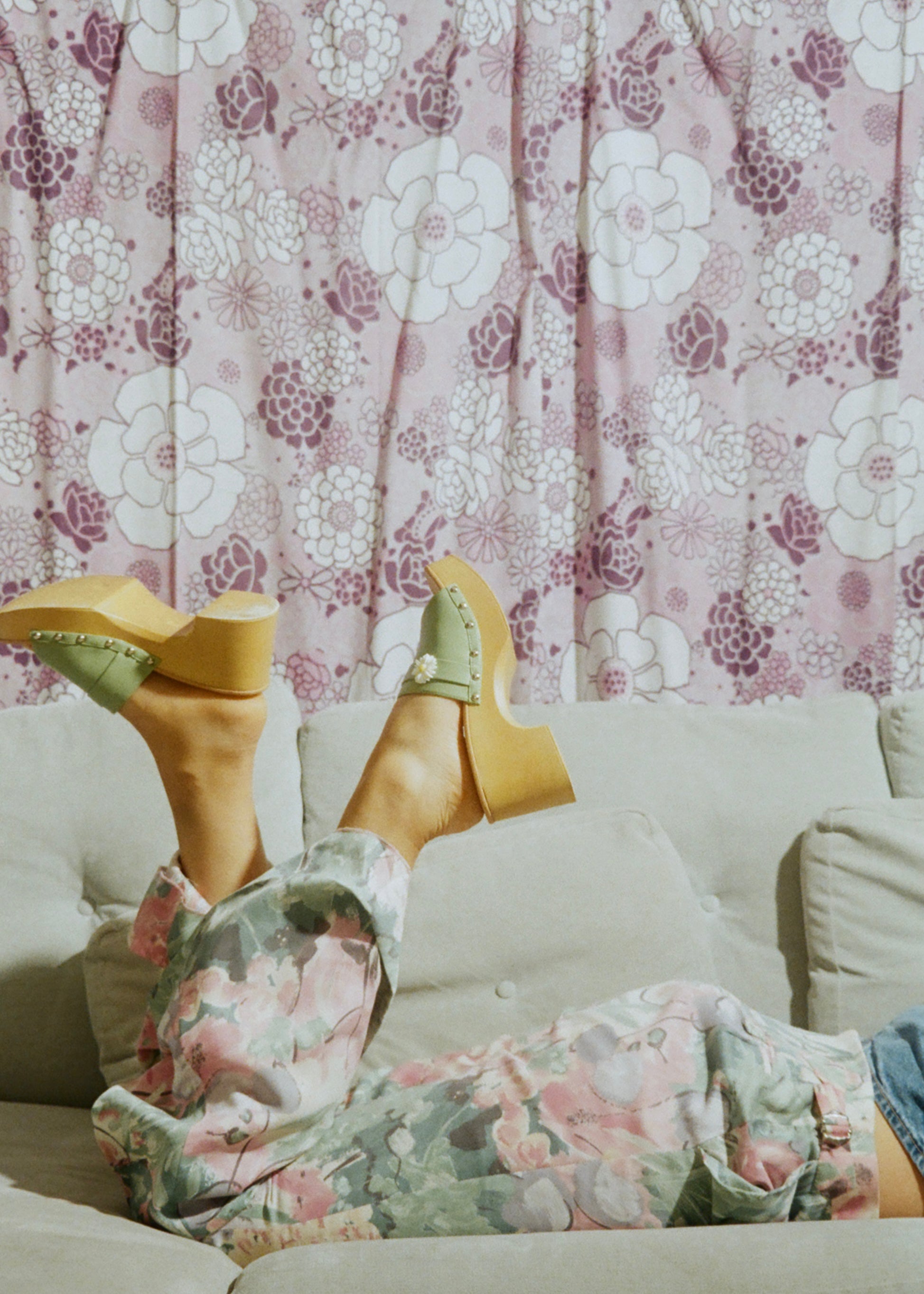 Person wearing floral pants and yellow high-heeled shoes with a floral curtain background