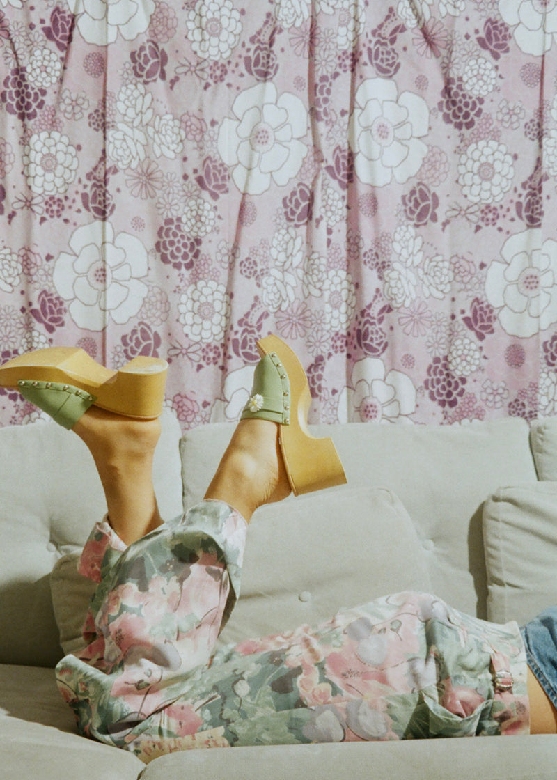 Person wearing floral pants and yellow high-heeled shoes with a floral curtain background