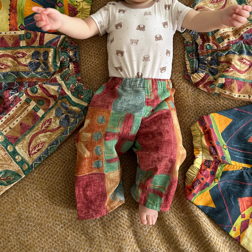 Child wearing patterned pants and a white shirt with animal designs, standing on a textured surface with colorful fabric pieces.