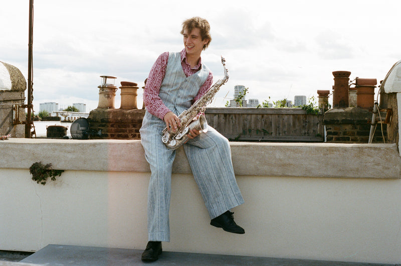 Person sitting on a rooftop with a saxophone, wearing a checkered shirt and overalls.