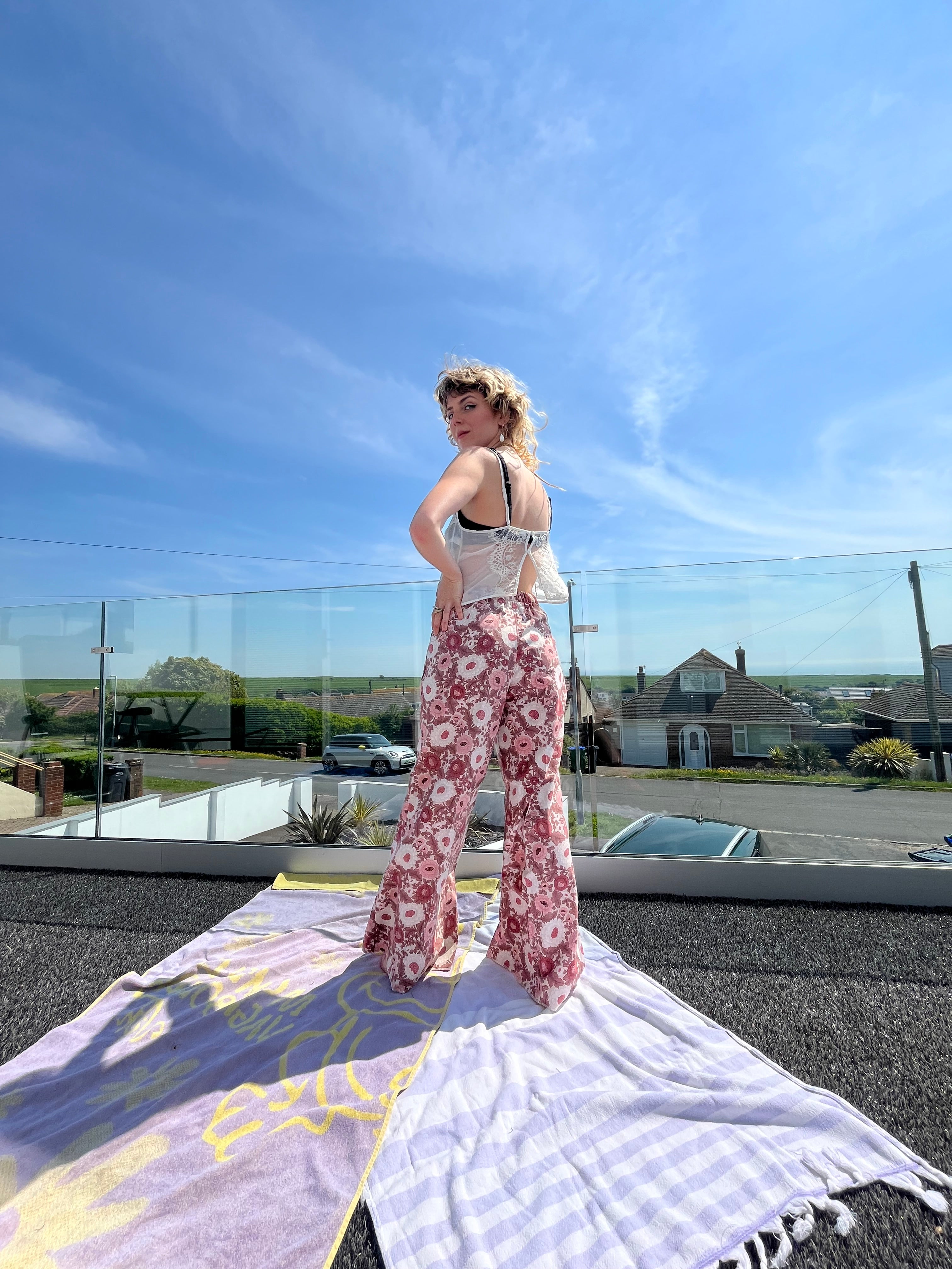 Person wearing a white tank top and floral pants standing on a balcony with a clear blue sky.