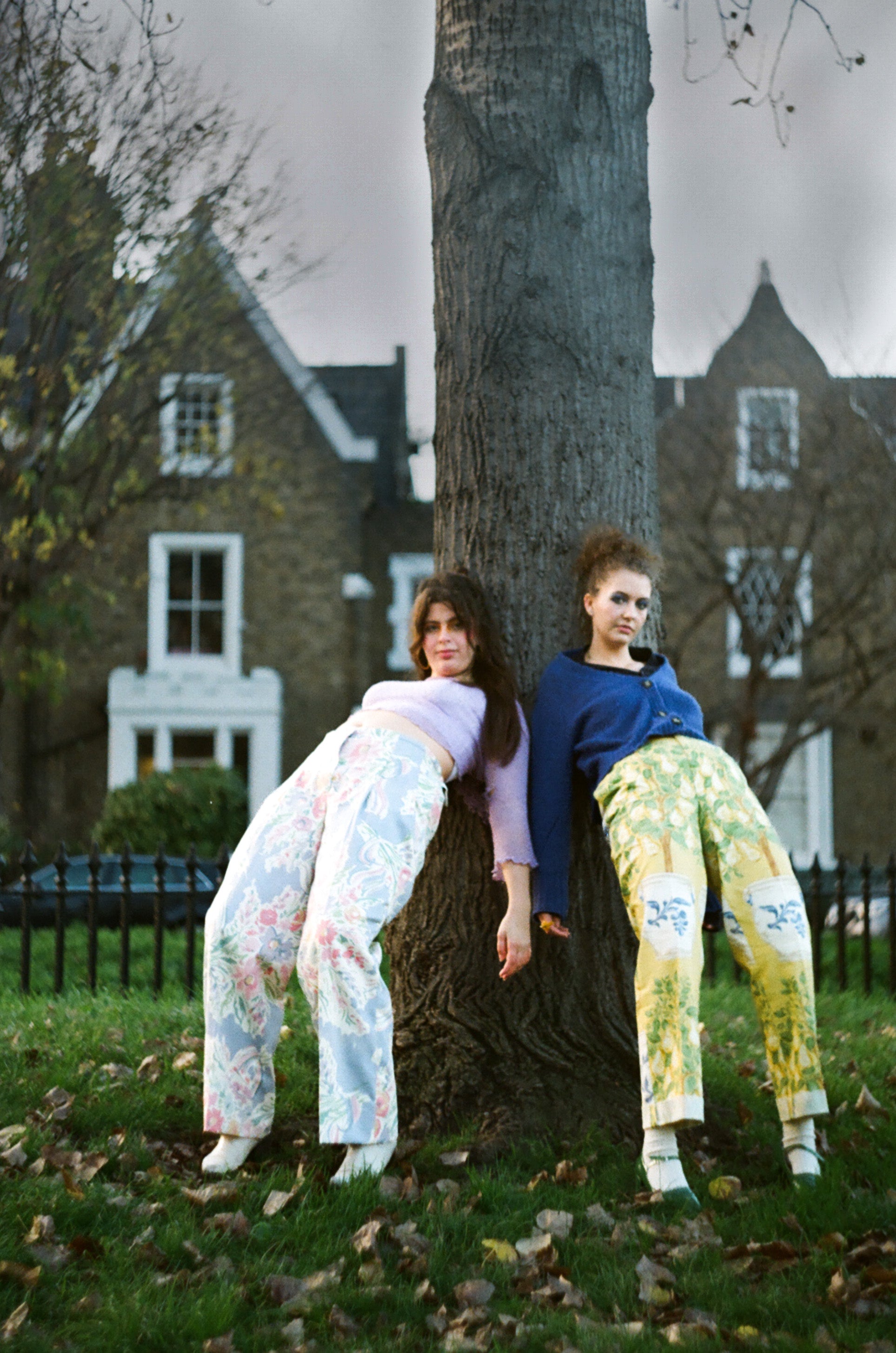 Two women leaning against a tree in a park with houses in the background