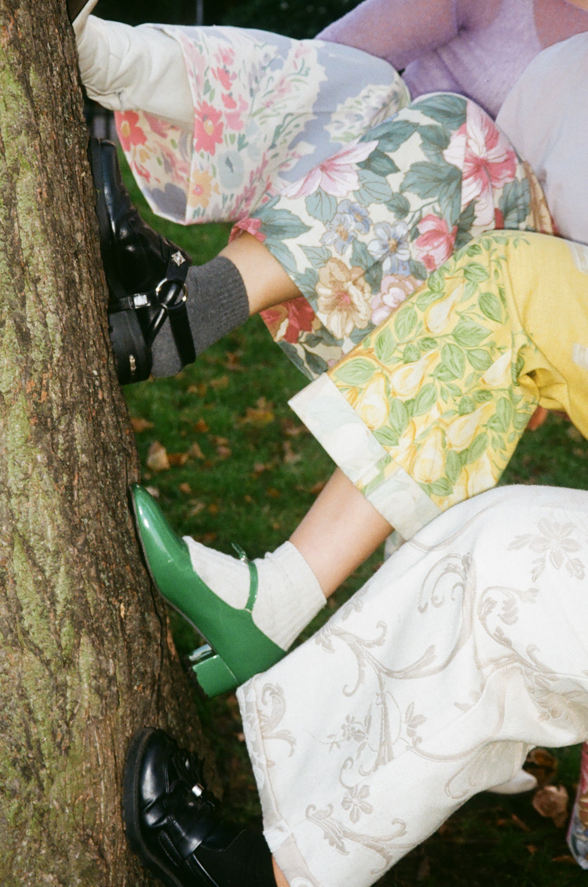 Person wearing floral shorts, white shoes, and green boots leaning against a tree.
